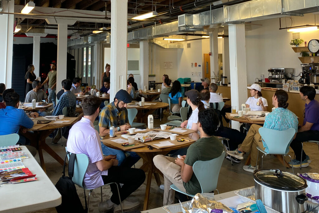 A large work team, dressed in casual attire for an offsite at the Lower Manhattan Cultural Council Art Center, is seated at several circular tables in a large room. Resting on the tables are painting supplies and worksheets for team activities.