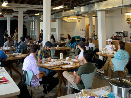 A large work team, dressed in casual attire for an offsite at the Lower Manhattan Cultural Council Art Center, is seated at several circular tables in a large room. Resting on the tables are painting supplies and worksheets for team activities.