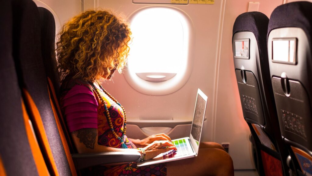 A woman living the lifestyle of a digital nomad is sitting on plane and doing work on a laptop in her lap.